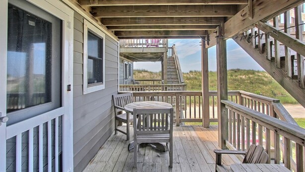 A wooden deck with a table and chairs, a staircase leading to a second level, and a view of the beach. - 27 Oceans Door East Studio Bedroom Condo by RedAwning (Hatteras)