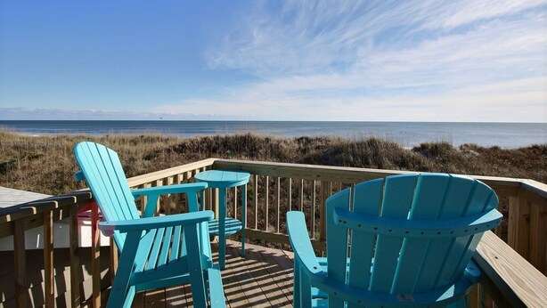 A wooden deck with blue Adirondack chairs overlooking a beach.