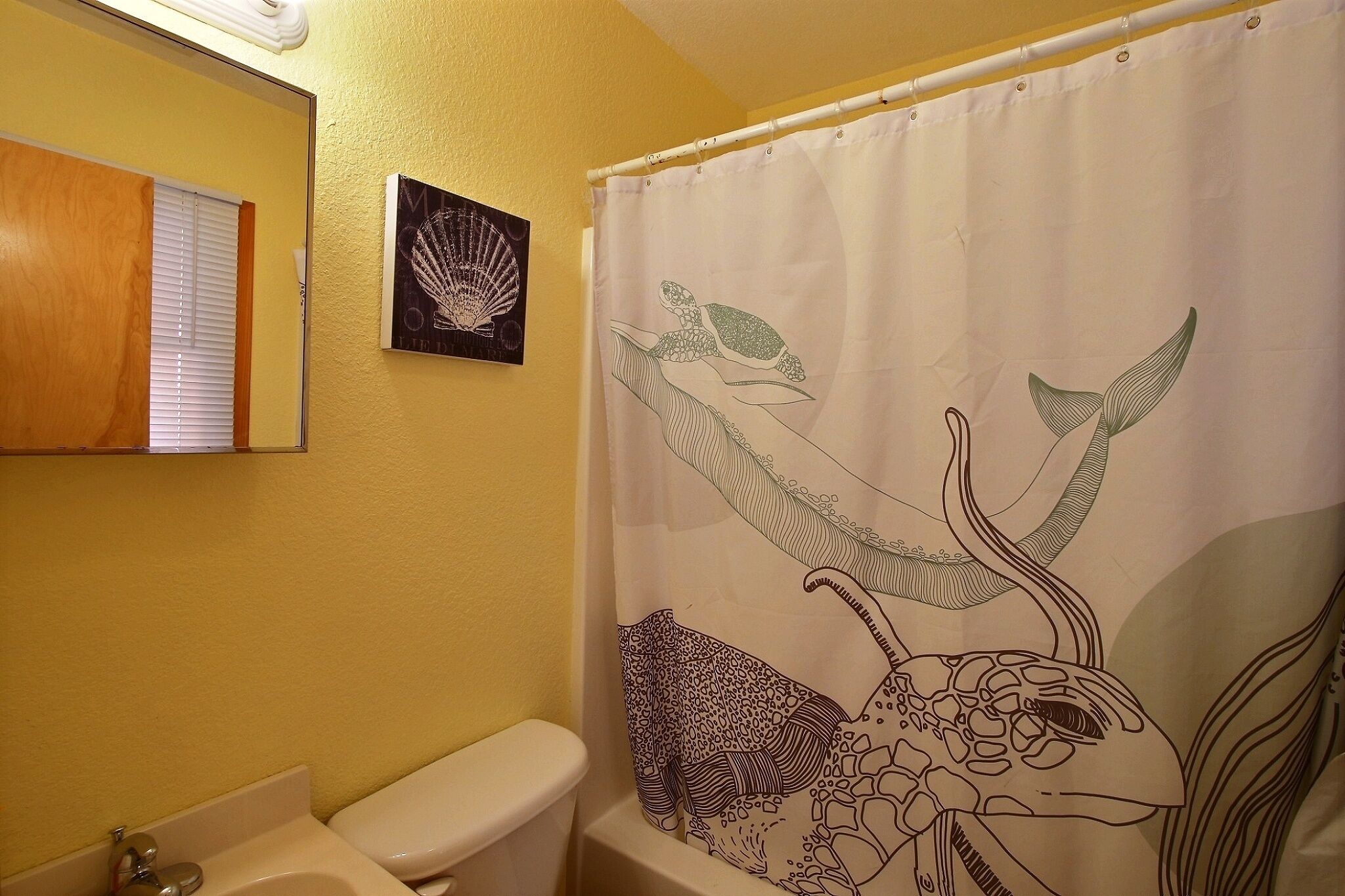 Un baño con pared amarilla, inodoro blanco, lavabo y cortina de ducha con diseño de tortuga.