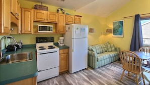 A compact kitchen with wooden cabinets, a microwave, a refrigerator, and a dining area with a striped sofa and wooden chairs.