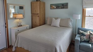 Une chambre à coucher comprenant un lit, une armoire en bois, une table de chevet, une lampe, un miroir, un ventilateur de plafond et une fenêtre avec des rideaux.