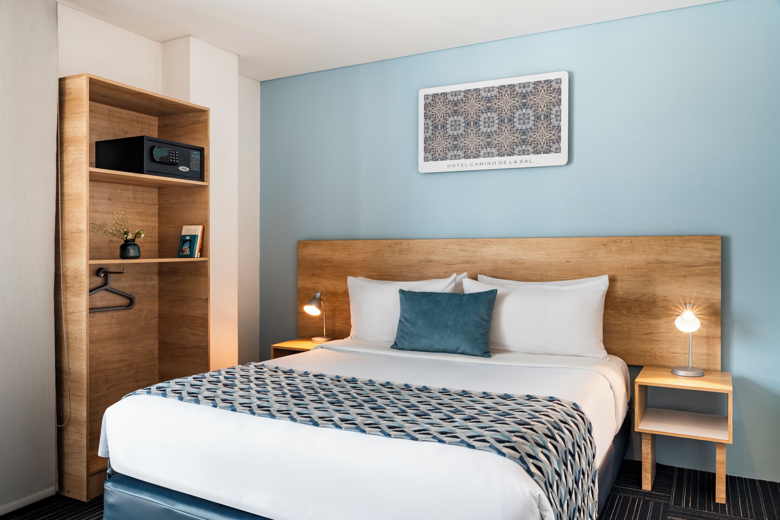 A hotel room with a wooden headboard, a bed with patterned bedding, a nightstand with a lamp, a wooden wardrobe, and a framed picture on the wall.