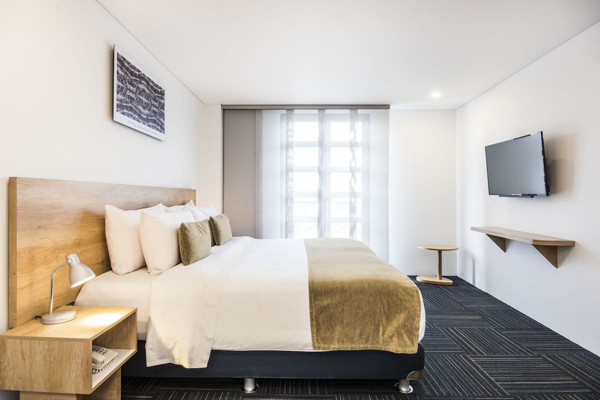 Une chambre d’hôtel avec un lit, une table de chevet, une lampe, une télévision et une fenêtre avec des rideaux.