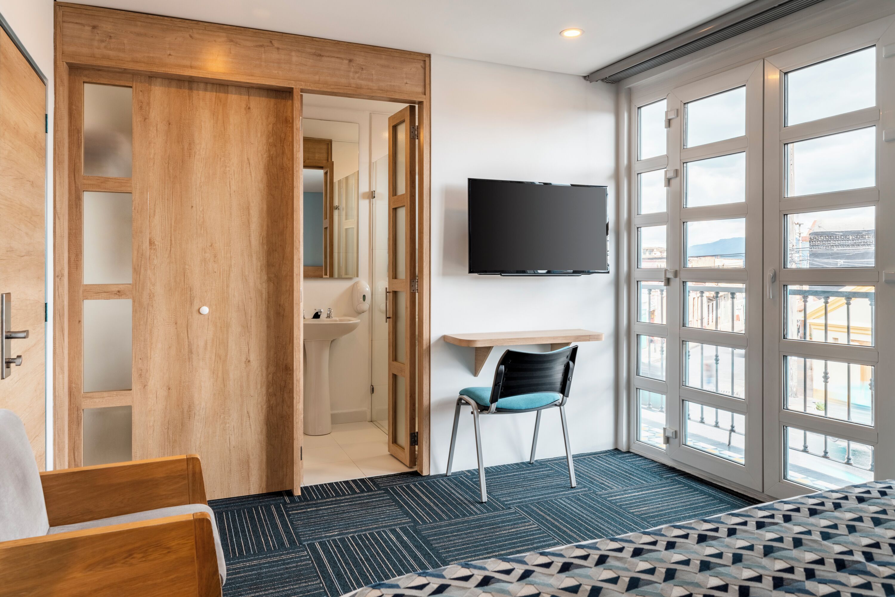 A hotel room with a bed, a desk, a chair, a TV, a bathroom, and large windows.