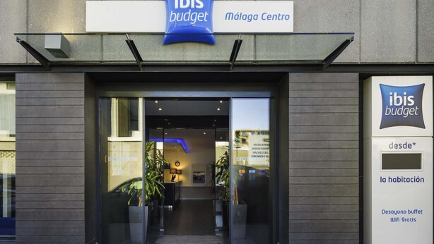 De ingang van een ibis budget hotel met een modern ontwerp, met een glazen deur, een bord en een digitaal scherm.