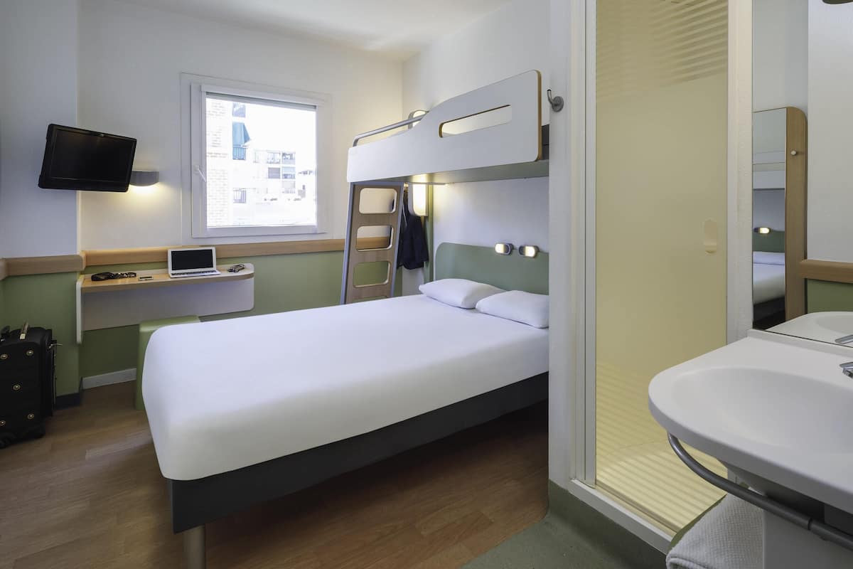 Une chambre d’hôtel de taille réduite, comprenant un lit simple, un bureau avec un ordinateur portable, un téléviseur fixé au mur et une salle de bain avec un lavabo et un miroir.