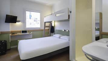 Habitación de hotel compacta con una cama individual, un escritorio con una computadora portátil, un televisor montado en la pared y un baño con lavamanos y espejo.