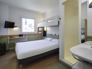 Une chambre d’hôtel de taille réduite, comprenant un lit simple, un bureau avec un ordinateur portable, un téléviseur fixé au mur et une salle de bain avec un lavabo et un miroir.