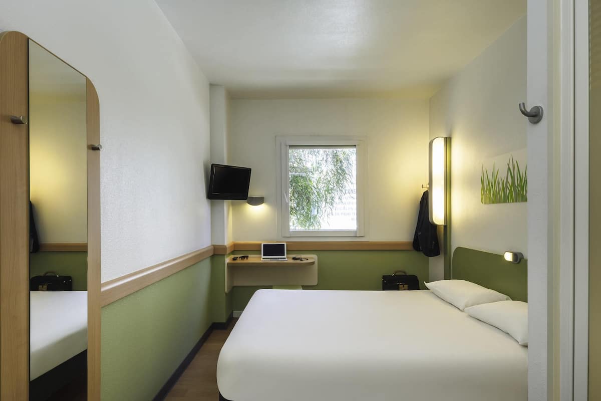 Une chambre d’hôtel avec un lit, un bureau avec un ordinateur portable, une télévision, une fenêtre et un miroir.