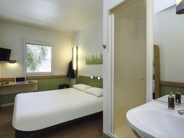 Habitación de hotel con cama, mesita de noche con una computadora portátil, ventana con persianas, televisor montado en la pared y baño con lavamanos y espejo.