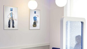Een moderne kamer met twee ingelijste kunstwerken, een blauwe bank, een witte tafel en een glazen scheidingswand, waarbij een persoon zichtbaar is.