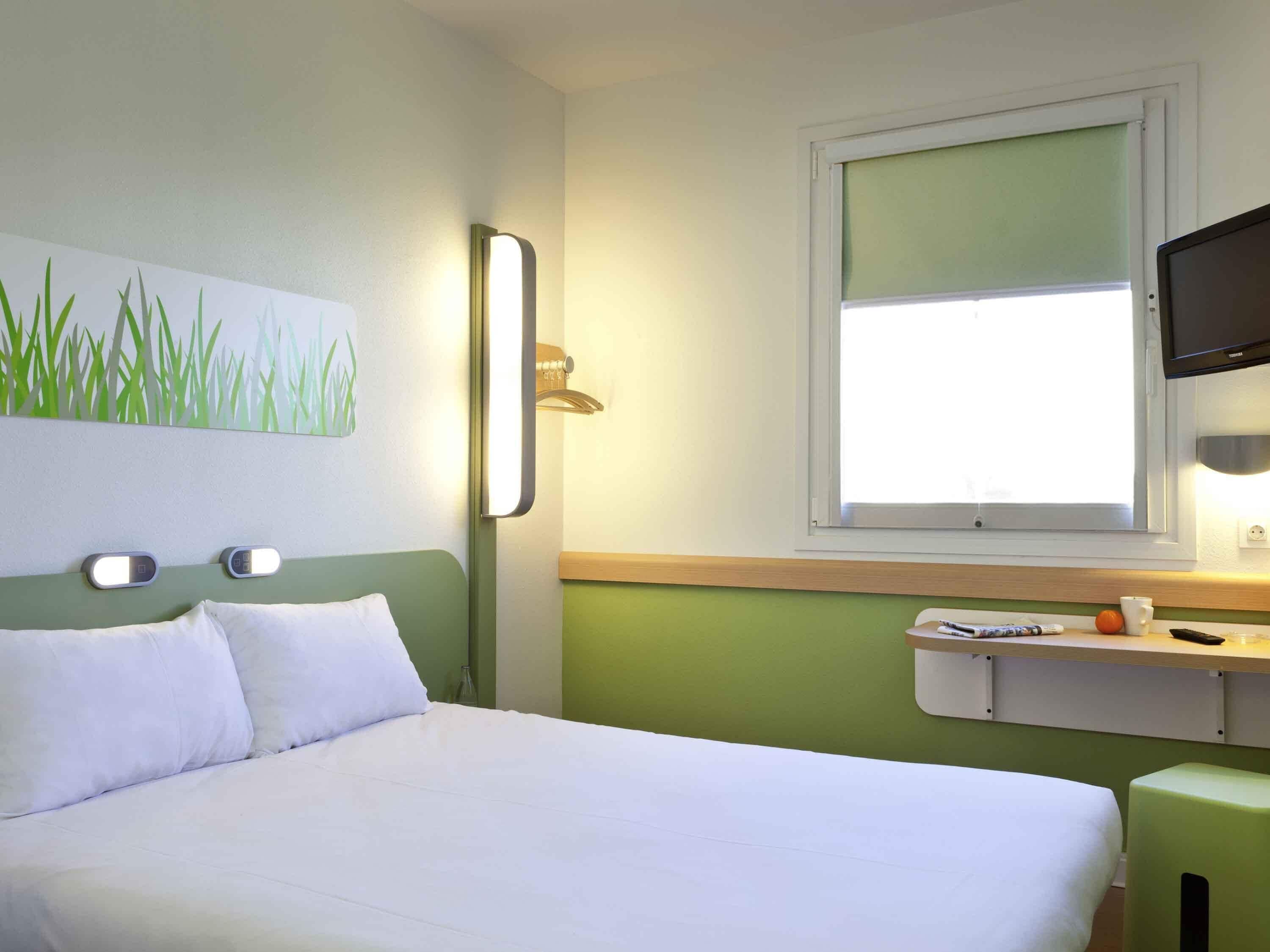 A hotel room with a bed, a painting of grass, a bedside table with a cup and a remote control, and a television mounted on the wall.