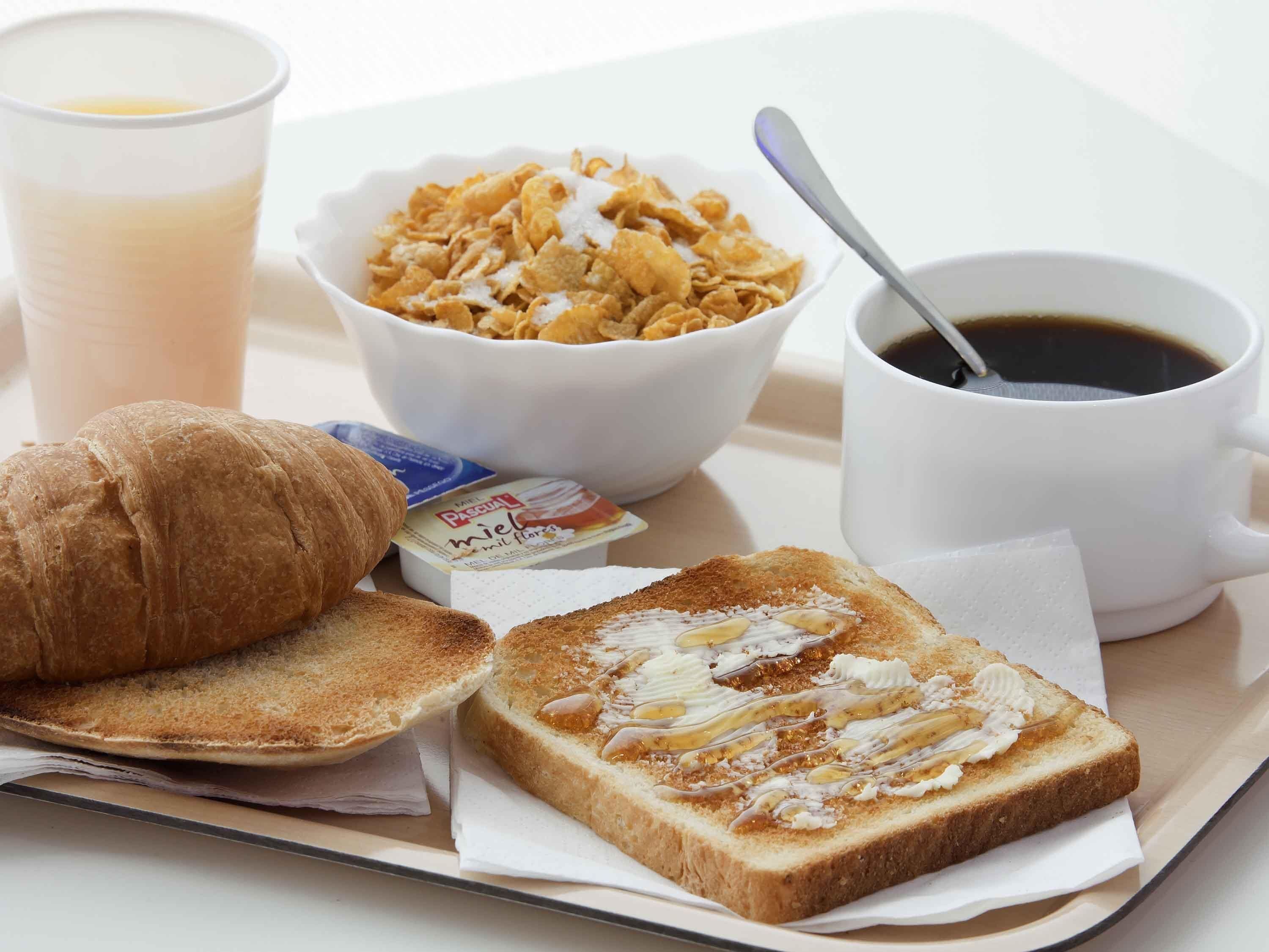 A breakfast tray with toast, cereal, a croissant, a cup of coffee, and a small container of milk.