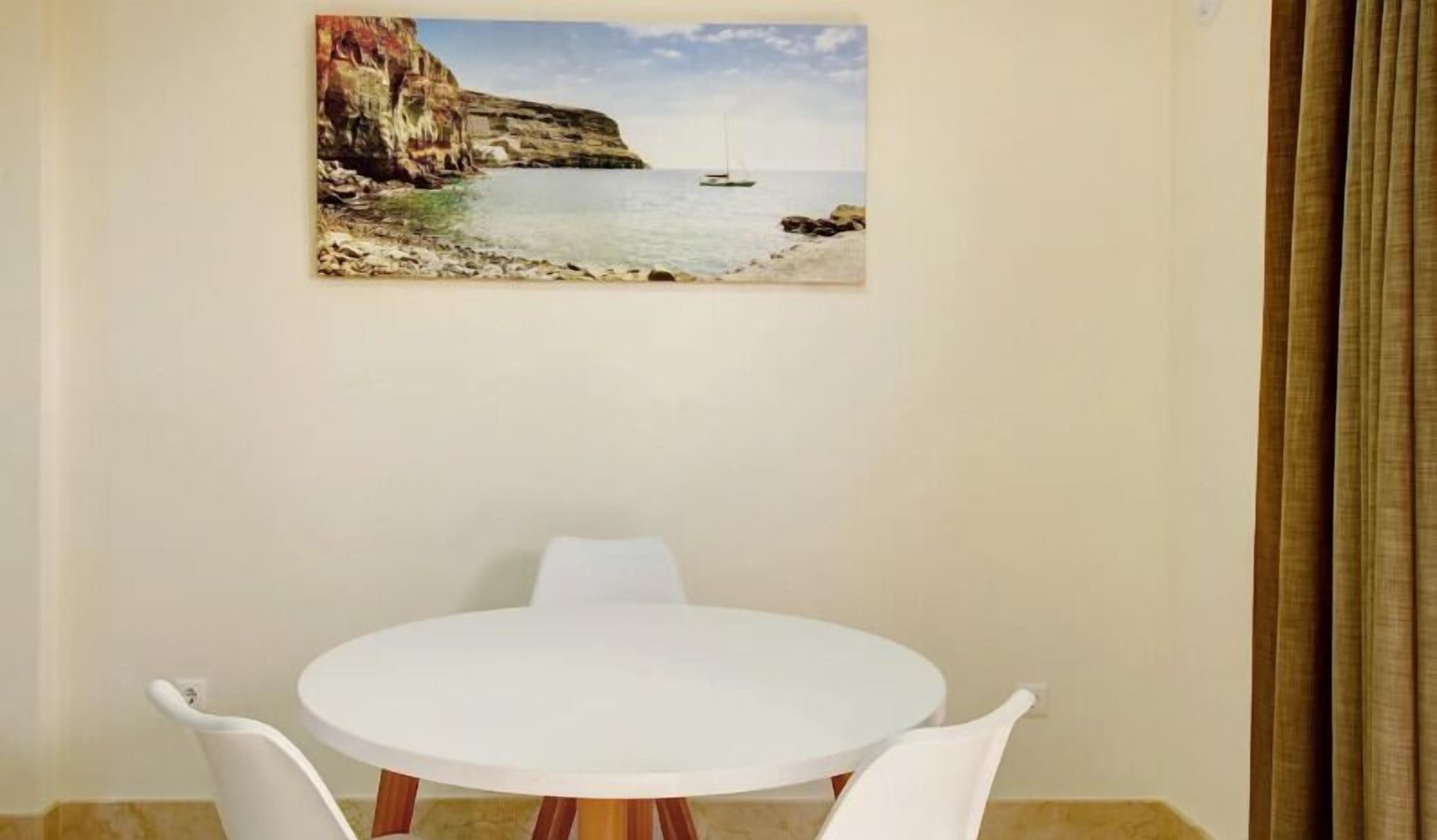A dining area with a round white table and two white chairs, a painting of a coastal scene on the wall, and a curtain on the right.