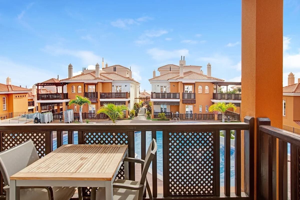 A balcony with a wooden table and chairs, overlooking a row of buildings with balconies and a clear sky.