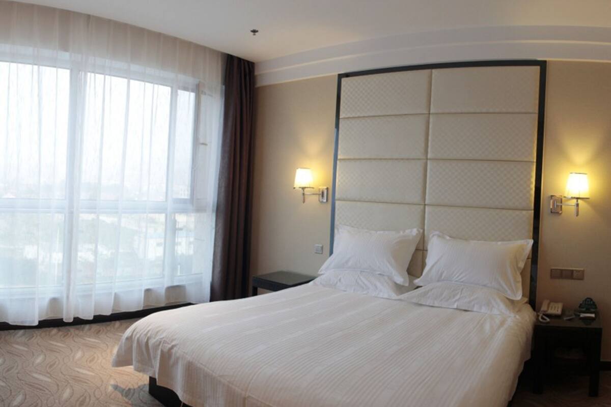 Una habitación de hotel con una cama grande, dos mesitas de noche, una lámpara de pared y una ventana con cortinas translúcidas.