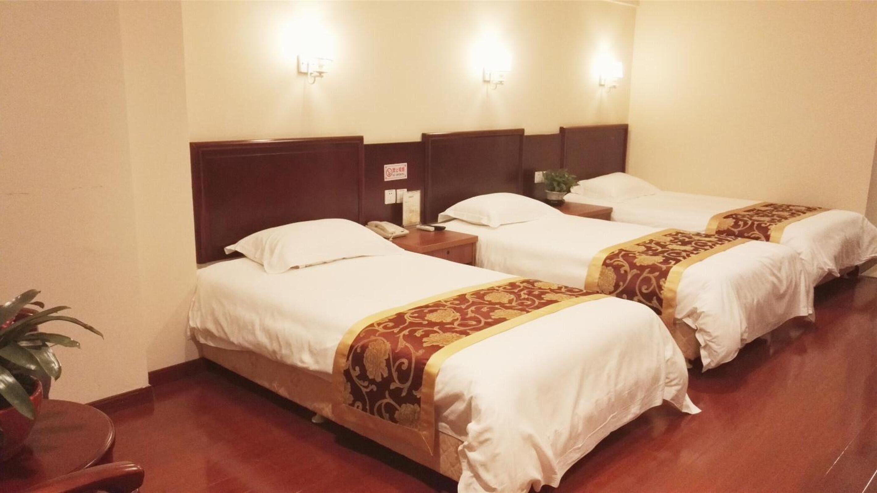 A hotel room with three single beds, each with a wooden headboard and white bedding. There is a small table with a telephone and a plant on it. The room has a wooden floor and beige walls.