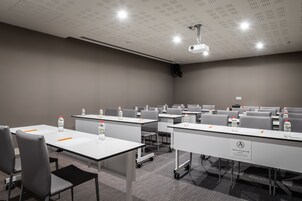 A conference room with tables, chairs, and a projector. Tables have water bottles and orange pens. The room has a gray carpet and a white ceiling with lights.