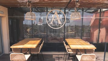 Een terras met houten tafels en rieten stoelen, een groot rond logo op de glazen wand, en hangende rieten lampen.