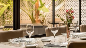 Una mesa de comedor preparada con cristalería transparente, platos y un jarrón con flores, con vista a follaje a través de amplias ventanas.