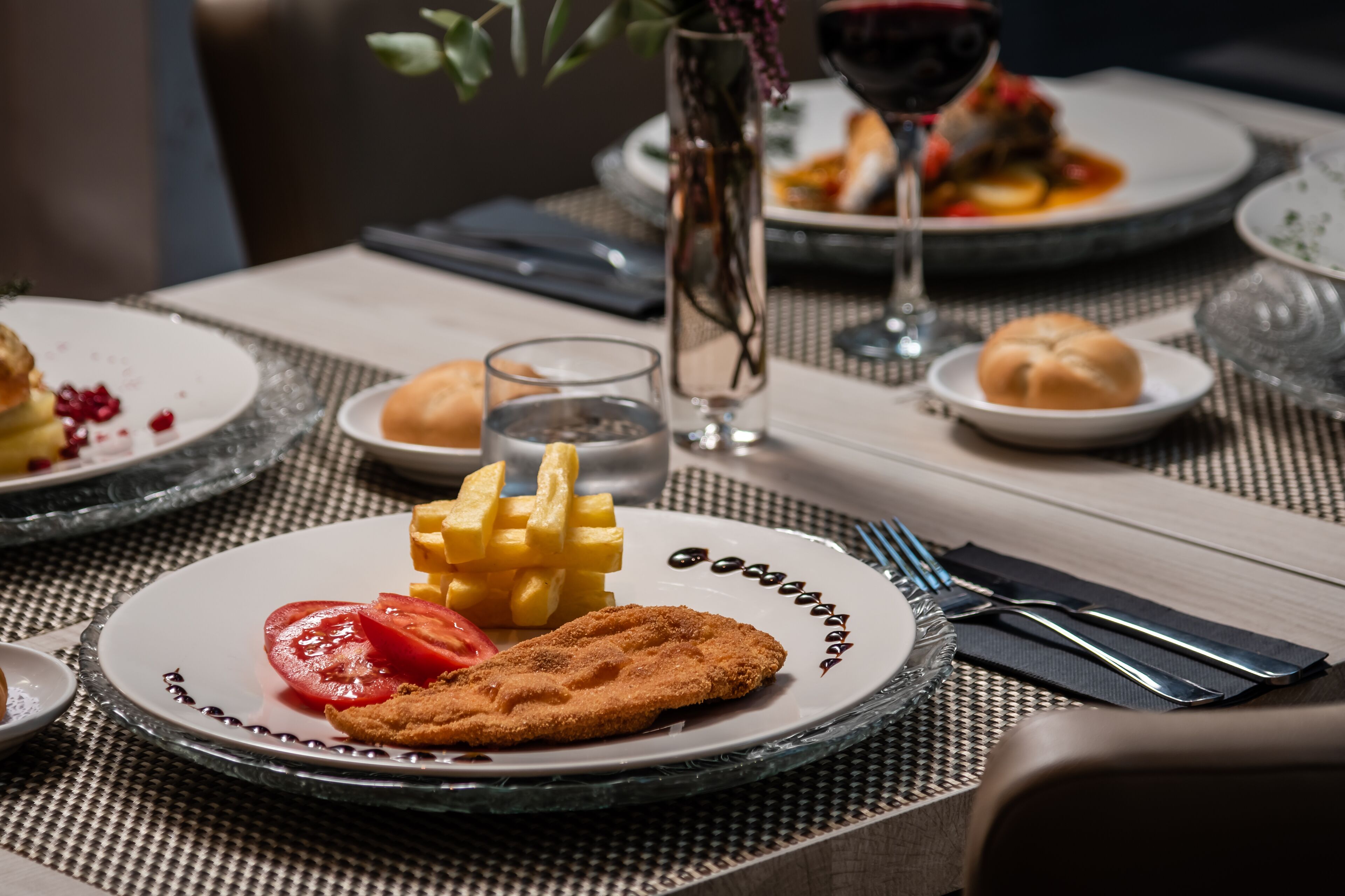 A dining table set with plates of food, including a breaded cutlet, fries, and a tomato slice.