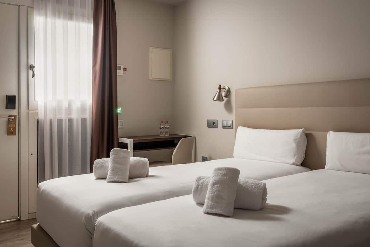 A hotel room with two beds, a desk, a chair, and a window with curtains.