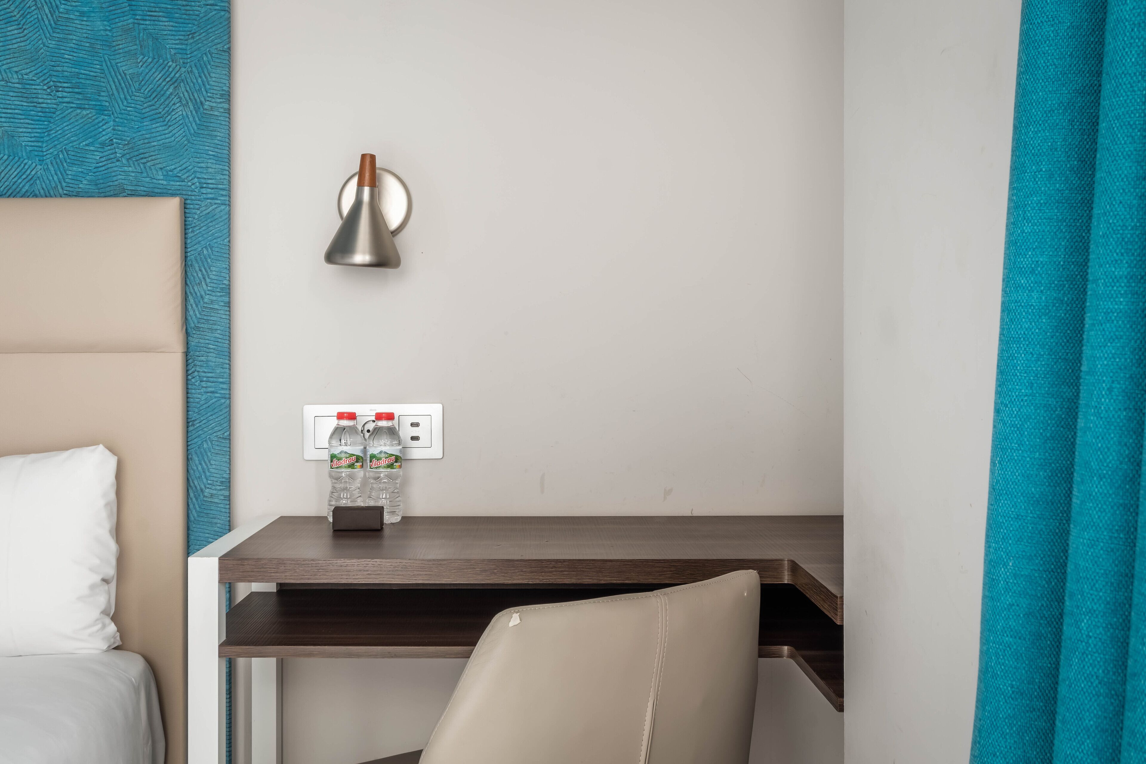 A hotel room with a bed, a desk, a lamp, and a bottle of water on the wall.