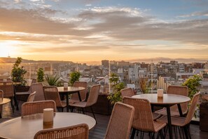 A rooftop terrace with wicker furniture and potted plants, overlooking a cityscape during sunset.