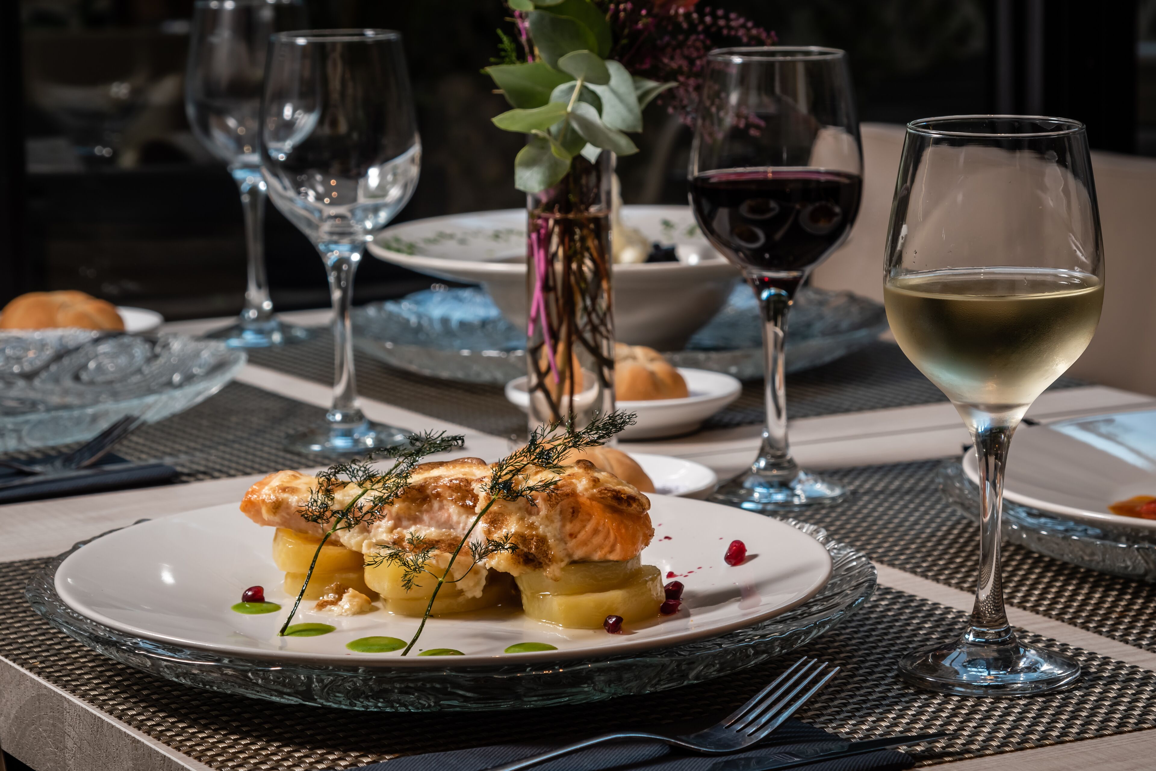 Una mesa de comedor con un plato de comida, copas de vino y un florero con plantas.