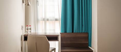 A hotel room with a bed, a desk with a chair, a bottle of water, and a window with curtains.
