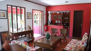 Un salón con muebles de mimbre, una mesa de centro de vidrio y un sofá con tapicería floral. Se observa una estantería con cuadros enmarcados y una puerta que da al exterior.