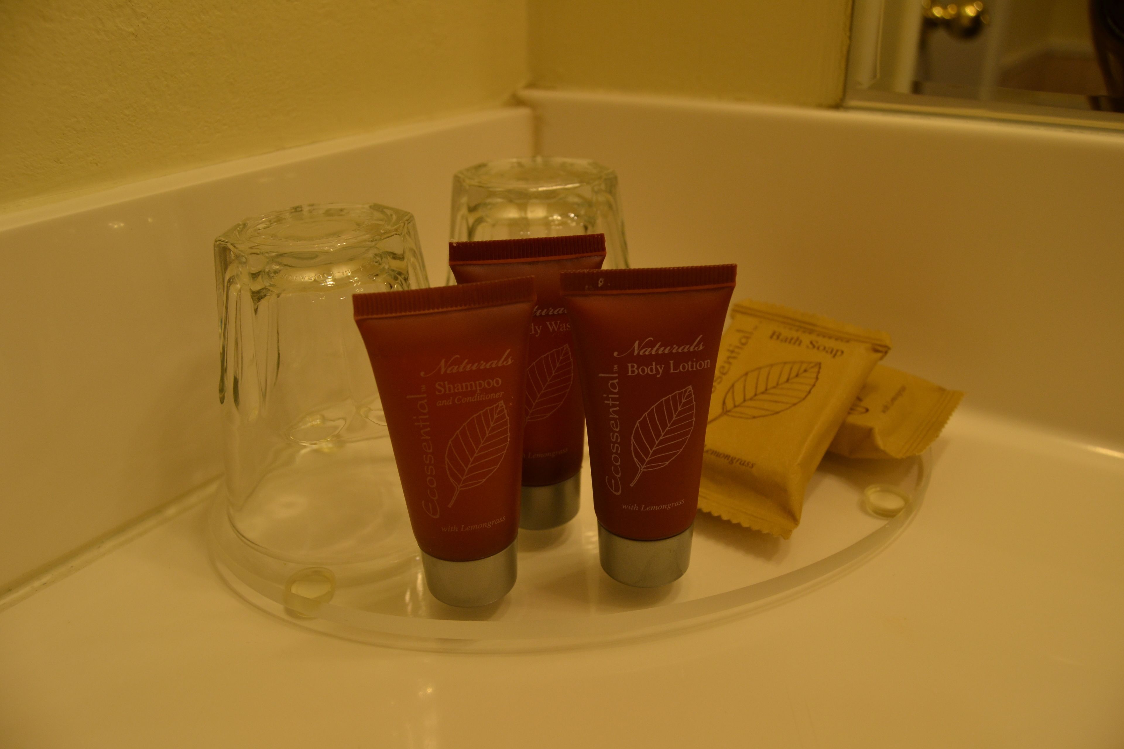 Uma prateleira no banheiro com produtos de higiene, incluindo um copo, dois tubos vermelhos de shampoo e condicionador, e uma pequena caixa marrom.