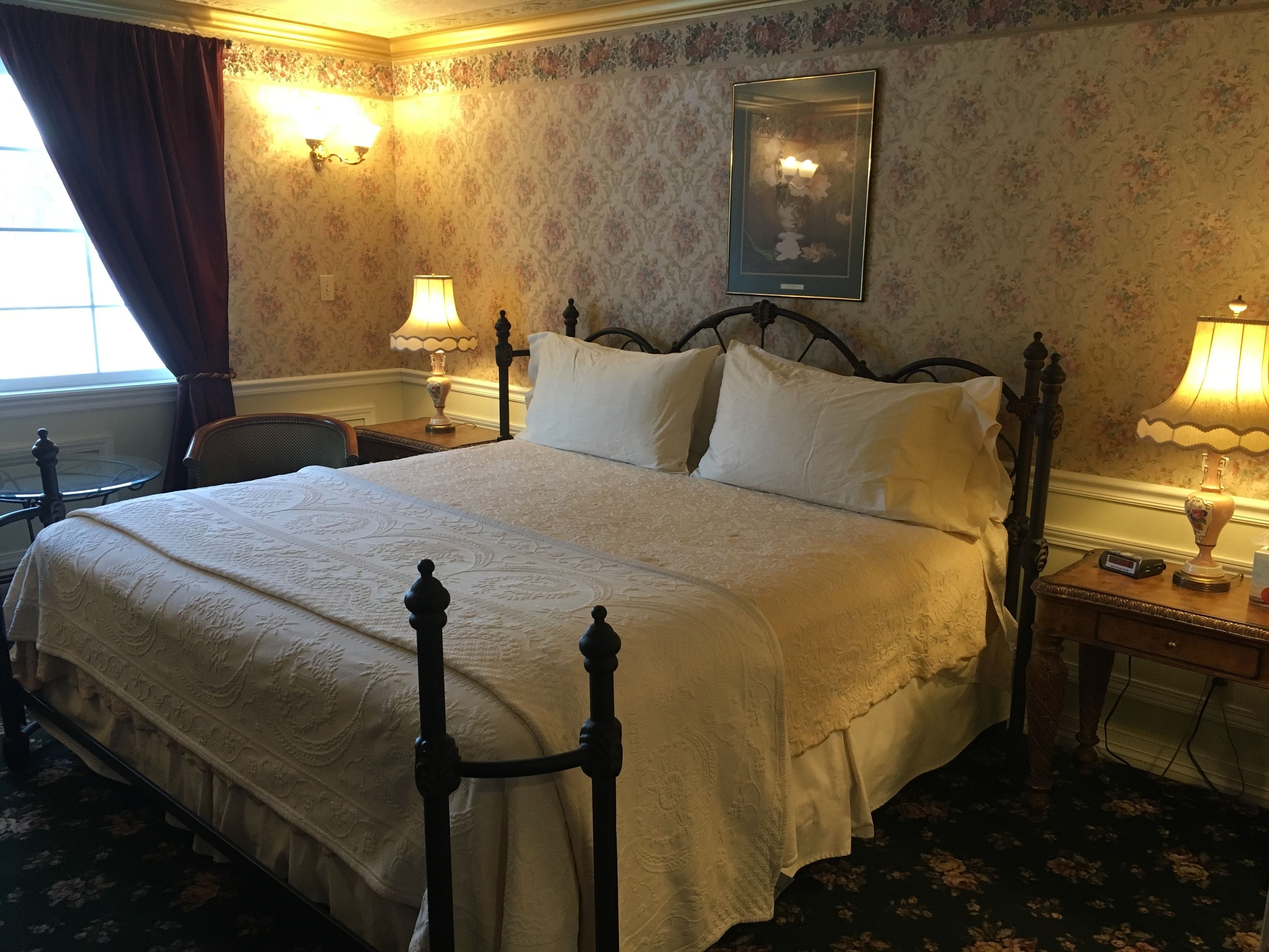 Una cama con dosel tradicional, con cubrecama y almohadas blancas, flanqueada por dos mesitas de noche con lámparas, en una habitación con papel tapiz floral y un cuadro enmarcado sobre la cama.