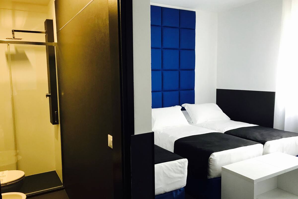 Habitación de hotel moderna con una paleta de colores blanco y negro, una cama grande con cabecera azul, una mesita de noche pequeña y un área de baño separada.