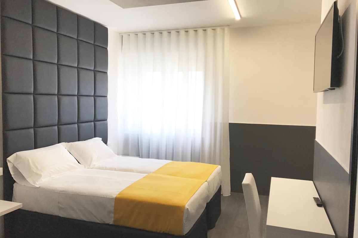 Habitación de hotel con cama, mesita de noche, televisor montado en la pared y ventana con cortinas.