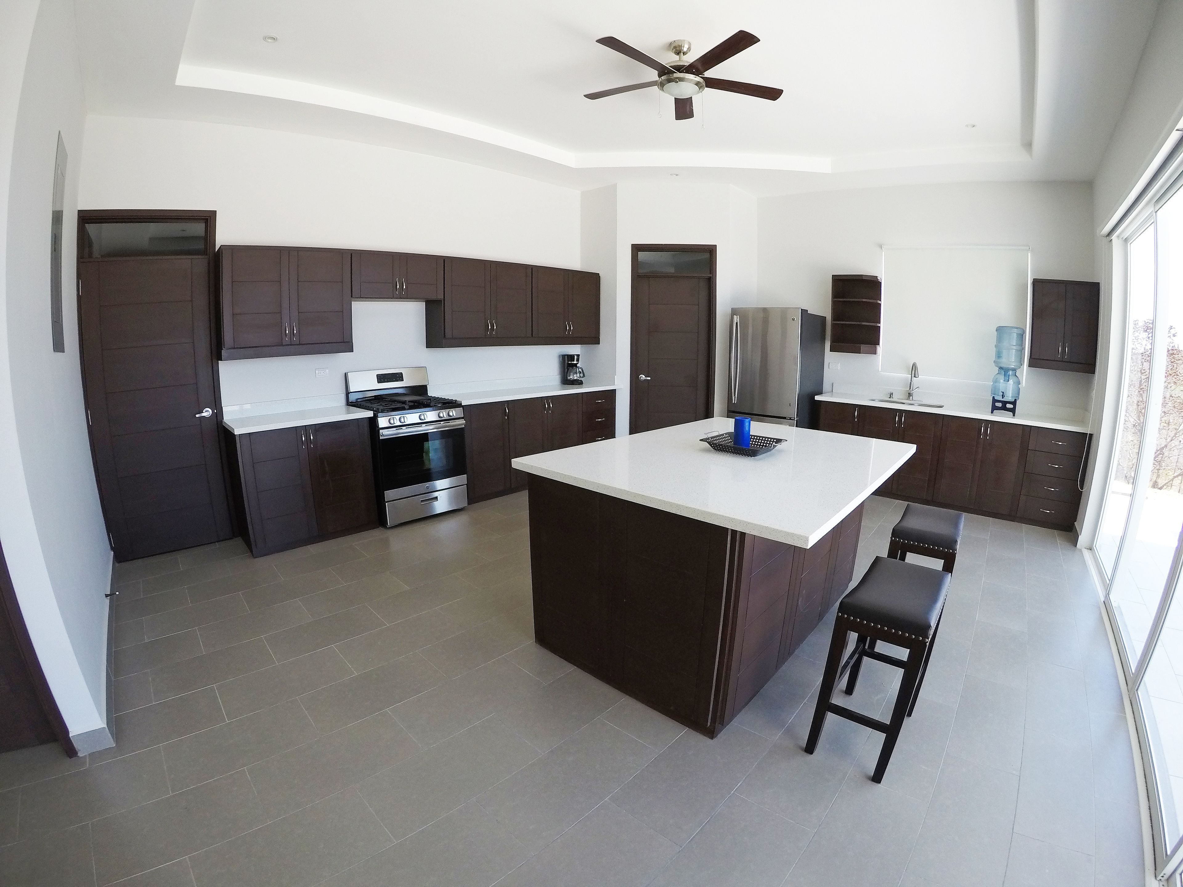 A modern kitchen with dark cabinetry, a central island, a built-in oven, and a ceiling fan.