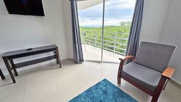A room with a large sliding glass door leading to a balcony, a wooden bench, a grey armchair, and a blue rug.