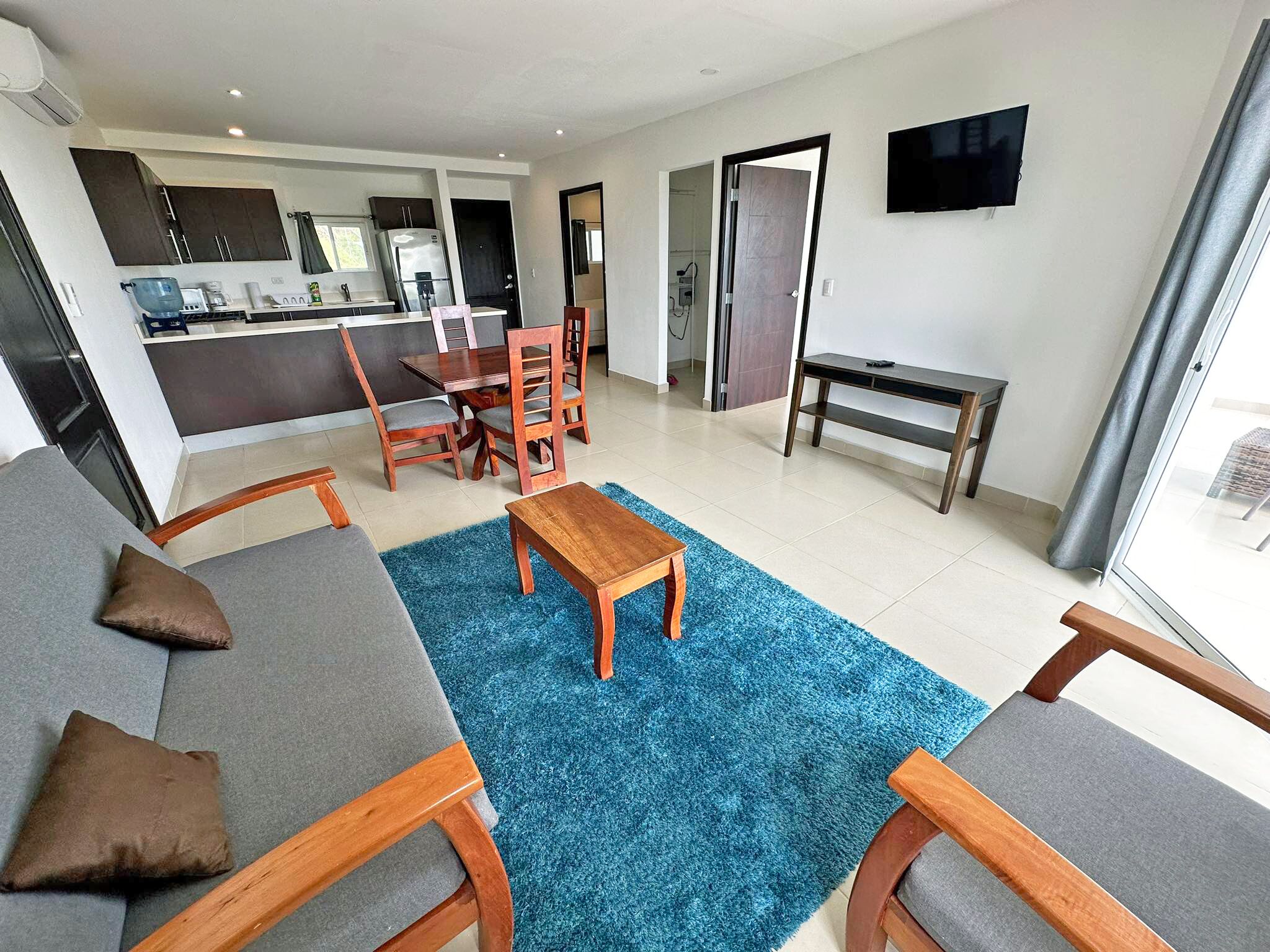 Un salon moderne avec un canapé gris, une table basse en bois et un tapis bleu. On y trouve également un coin cuisine avec un réfrigérateur et une table à manger avec des chaises. Un téléviseur est fixé au mur, et un banc est placé contre le mur.