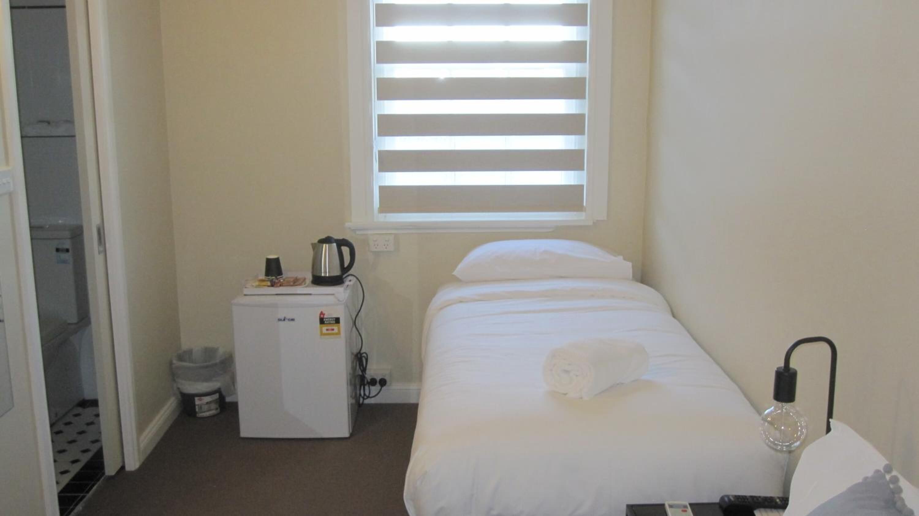 Quarto pequeno e limpo, com uma cama, um pequeno frigorífico, uma janela com persianas e uma lixeira.