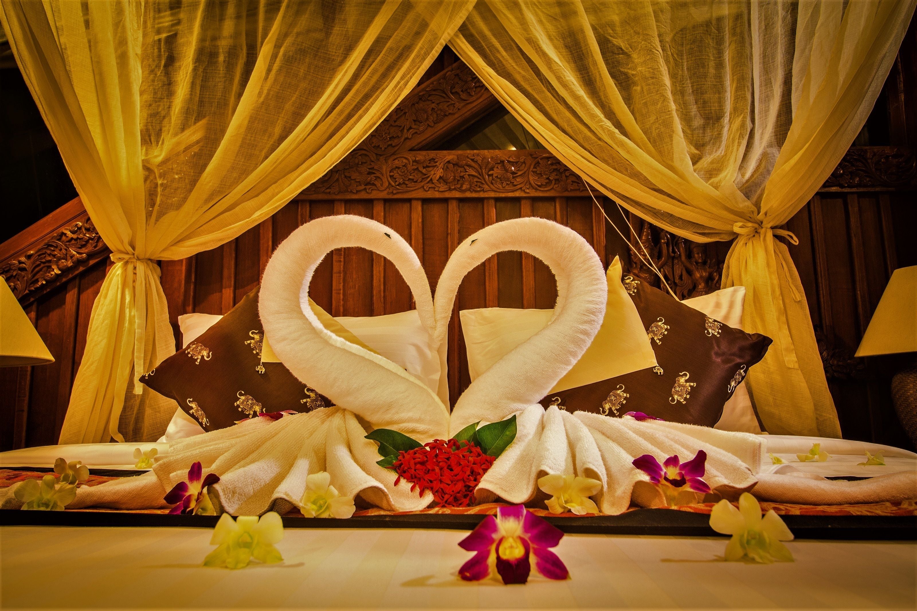A bed with a heart-shaped towel arrangement, a decorative pillow, and flowers.