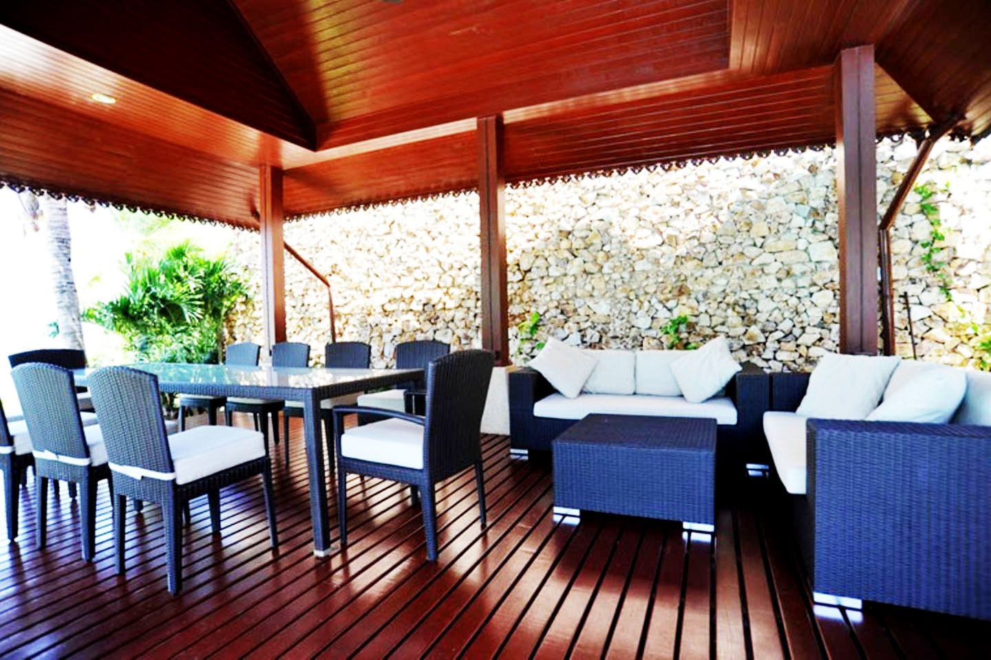 An outdoor covered patio with a stone wall, wooden flooring, and furniture including a table and chairs.