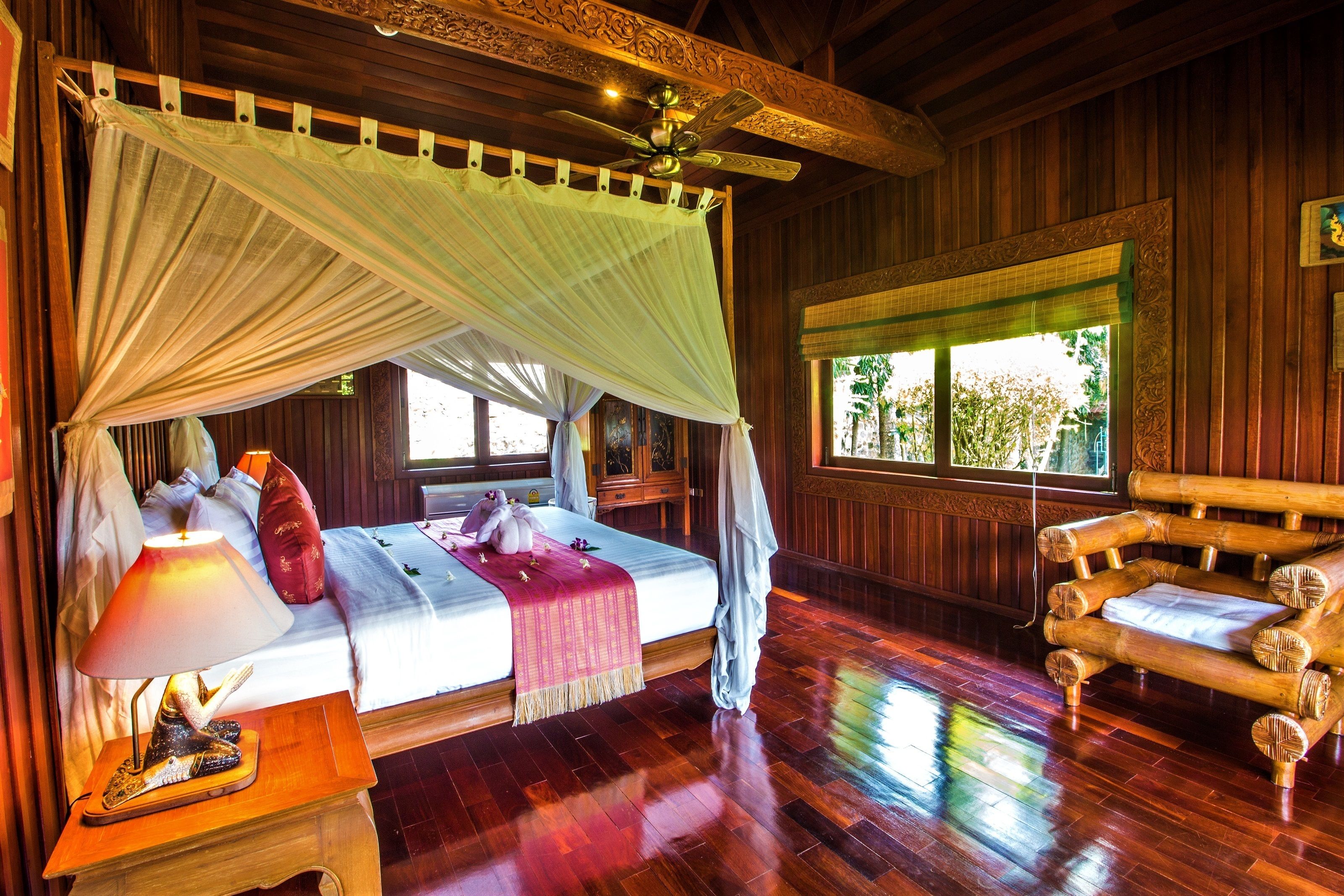 Une chambre avec un lit, une table de chevet, une chaise en bois, un ventilateur de plafond et une fenêtre donnant sur un paysage verdoyant.