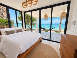 A bedroom with a large bed, a wooden dresser, and a view of the ocean through floor-to-ceiling windows.