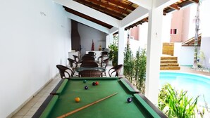 A pool table with balls, surrounded by wicker chairs, in a covered outdoor area with a pool in the background. - Pousada Portal do Sol (Bombinhas)