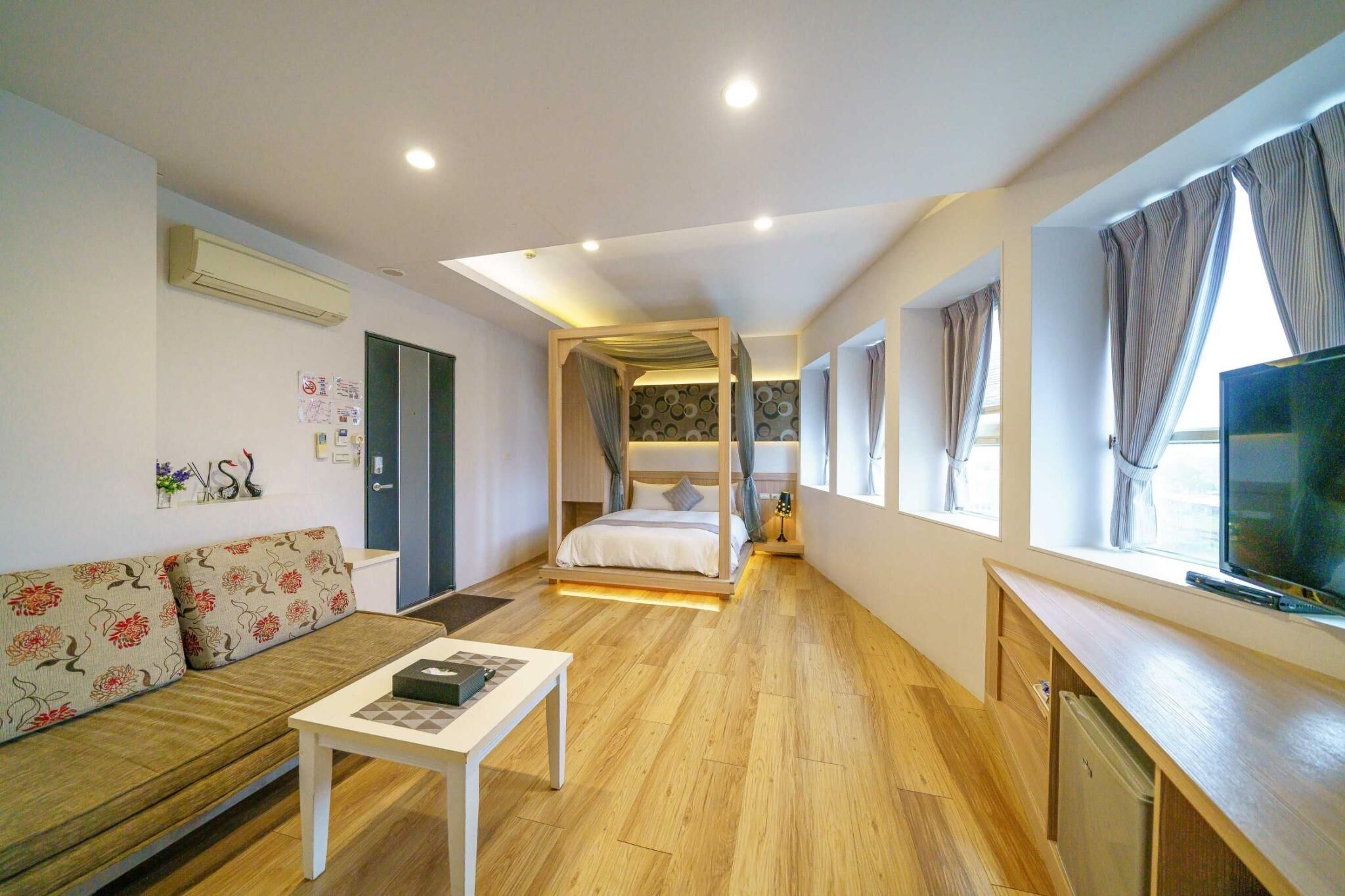 A modern hotel room with a wooden floor, a sofa, a bed with a canopy, a TV, and a desk.