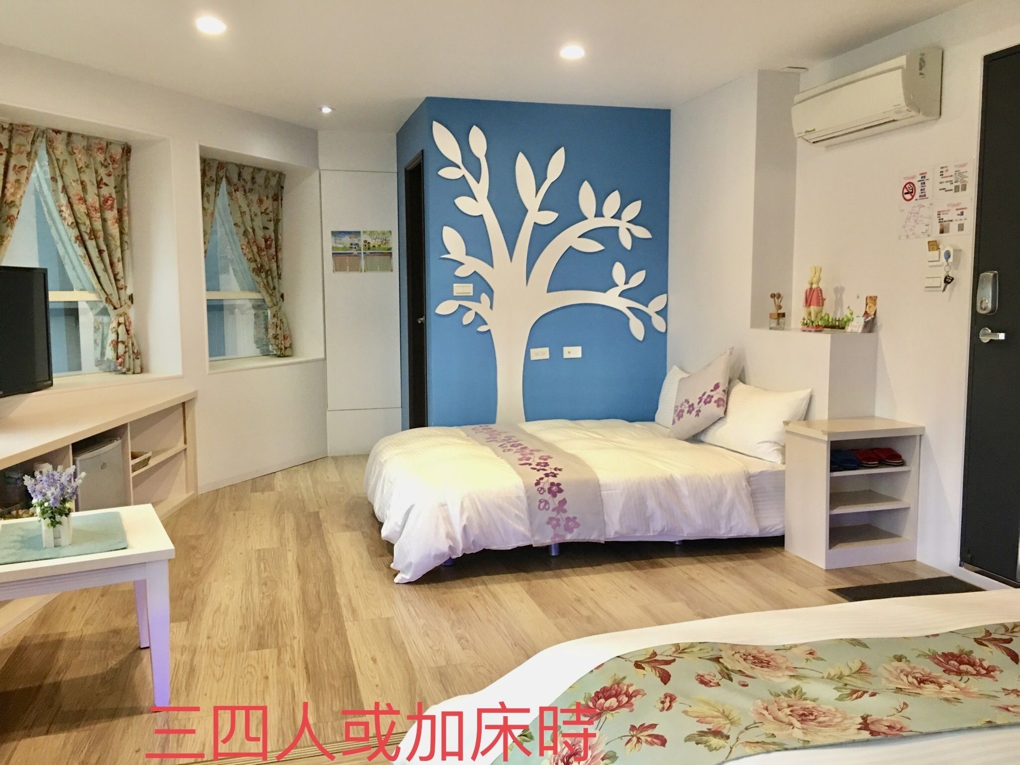 Ein Schlafzimmer mit Bett, Schreibtisch und Kleiderschrank. Eine blaue Wand zeigt ein Baumdesign, und an der Wand ist eine Klimaanlage angebracht.