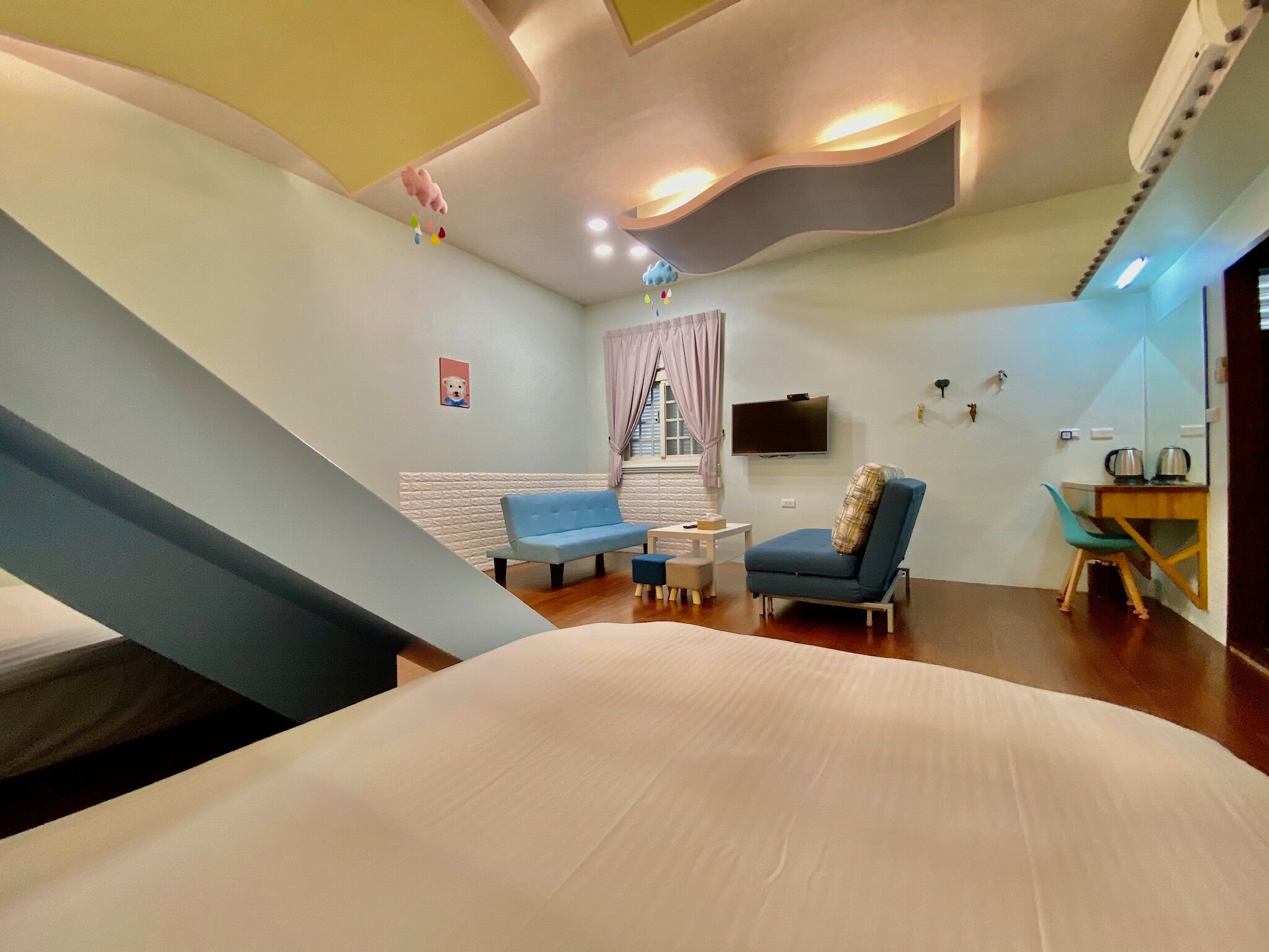 Una habitación de hotel moderna con una cama grande, un sofá azul, un sillón azul, una mesita y un televisor montado en la pared.