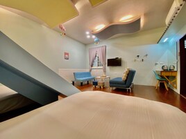 Una habitación de hotel moderna con una cama grande, un sofá azul, un sillón azul, una mesita y un televisor montado en la pared.
