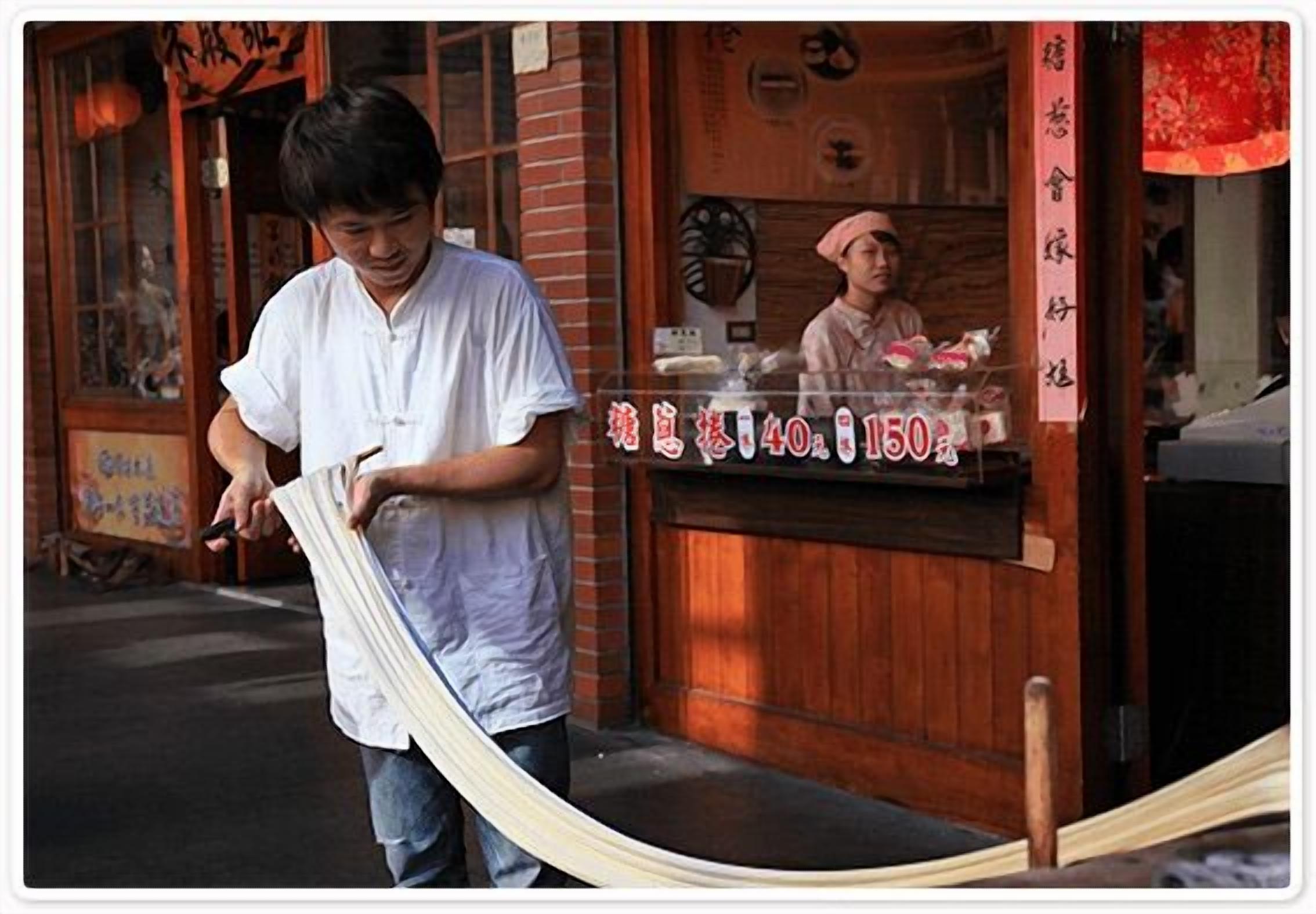 A person holding a long, light-colored fabric, standing in front of a building with a wooden facade and a sign with prices.