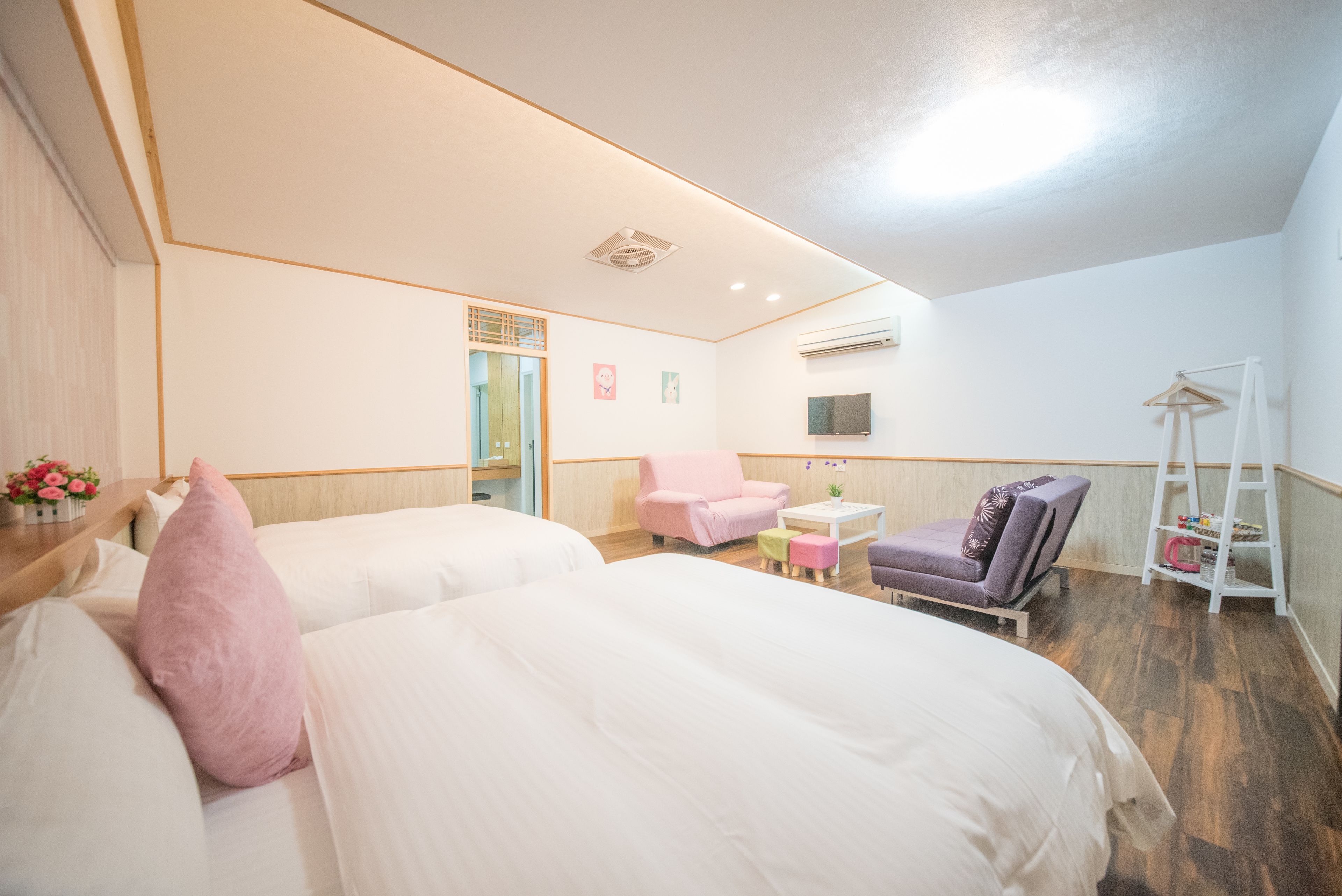 A hotel room with two beds, a pink chair, a purple chair, a small table, and a white shelf.